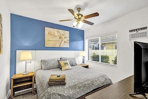 Une chambre à coucher comprenant un lit, deux tables de chevet, un ventilateur de plafond, une fenêtre avec stores et un climatiseur fixé au mur.