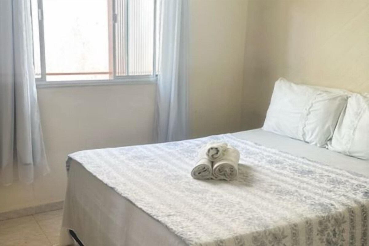 Un dormitorio con una cama, una ventana con cortinas y una toalla enrollada sobre la cama.