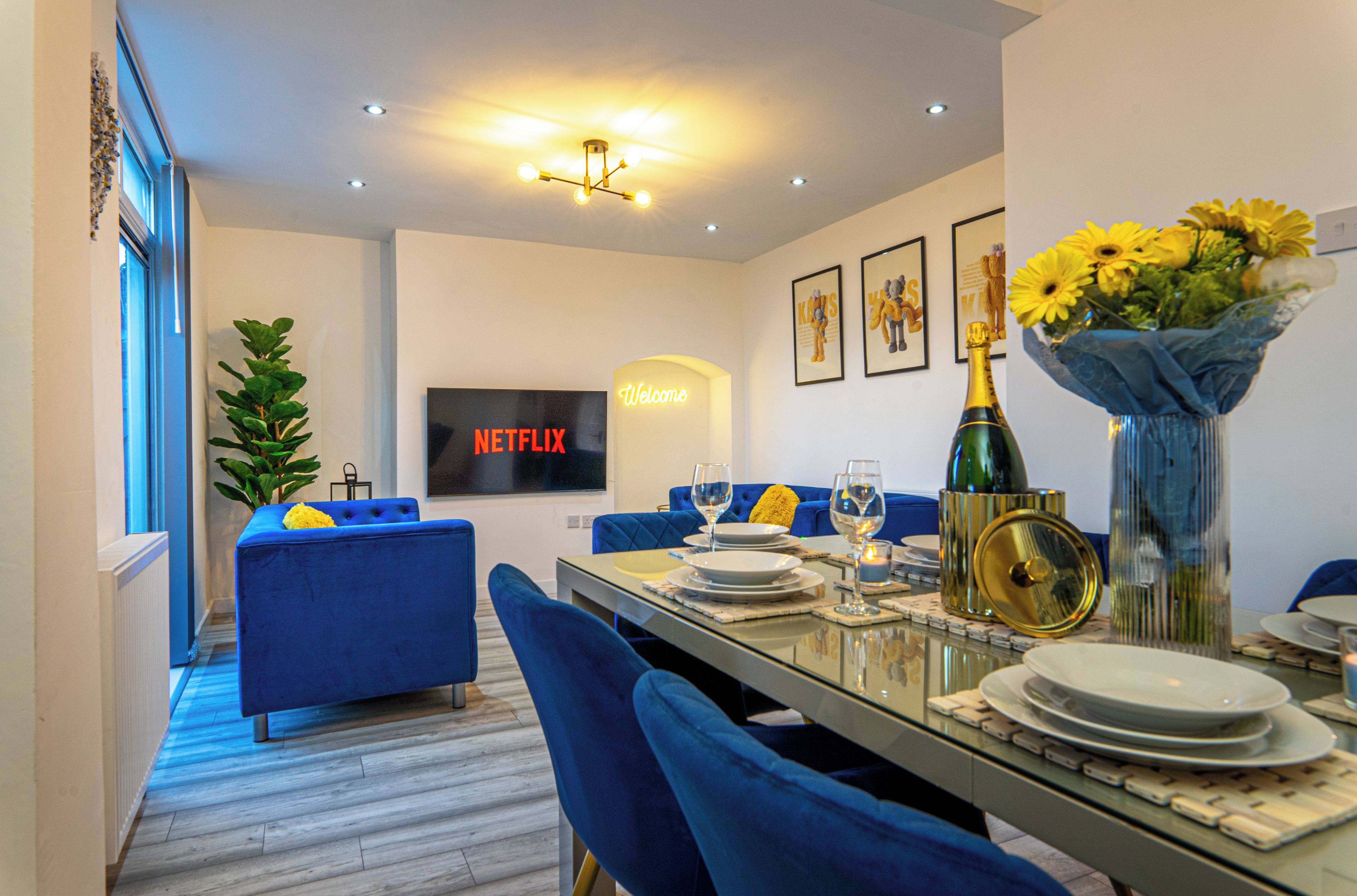 A modern dining area with a glass table, blue upholstered chairs, and a TV displaying "Netflix" in the background.