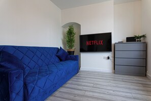 A room with a blue sofa, a television mounted on the wall, and a small plant on a cabinet. - Den Duplex | 3BR | Sleeps 6 | Free Parking (Rugby)