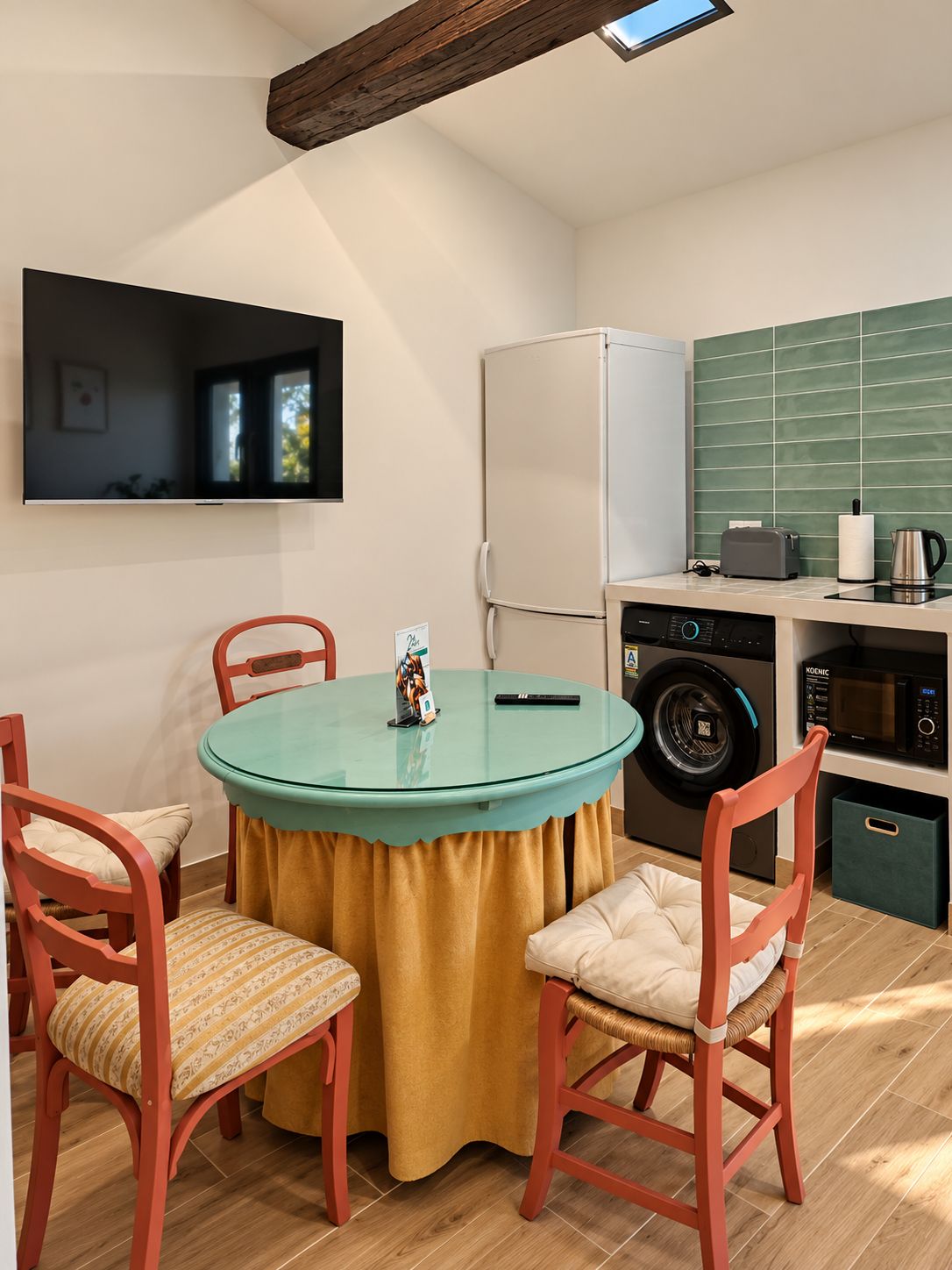 Cocina compacta con lavadora, microondas y refrigerador. Hay una mesa redonda con sillas y un televisor montado en la pared.