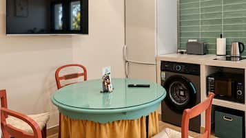 Cocina compacta con lavadora, microondas y refrigerador. Hay una mesa redonda con sillas y un televisor montado en la pared.