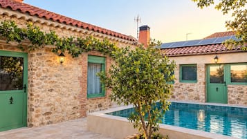 A stone building with a swimming pool, green doors, and a small tree in the foreground.
