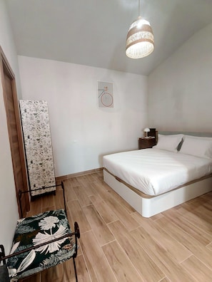 Ein Schlafzimmer mit einem Bett, einer Pendelleuchte, einem Wandbild und einem Sessel.