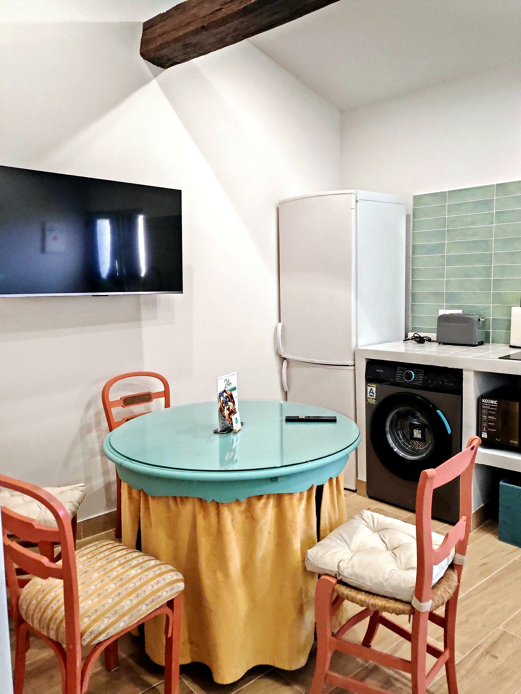 Una cocina pequeña con lavadora, refrigerador, una mesa con sillas y televisor montado en la pared.