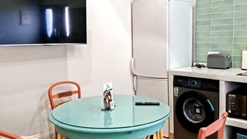 Una cocina pequeña con lavadora, refrigerador, una mesa con sillas y televisor montado en la pared.