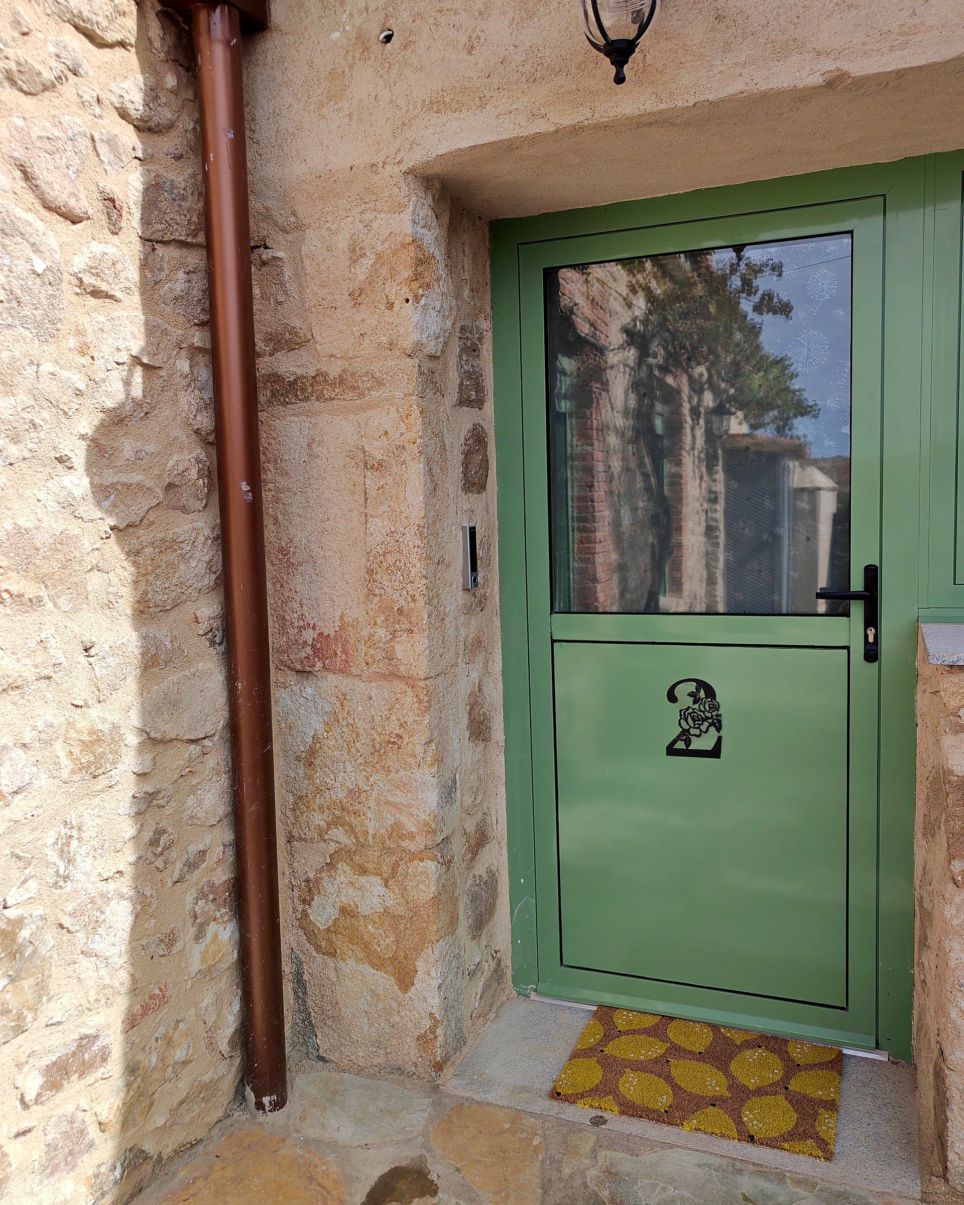 A green door with a number plate, a brown pipe, and a yellow mat at the entrance.