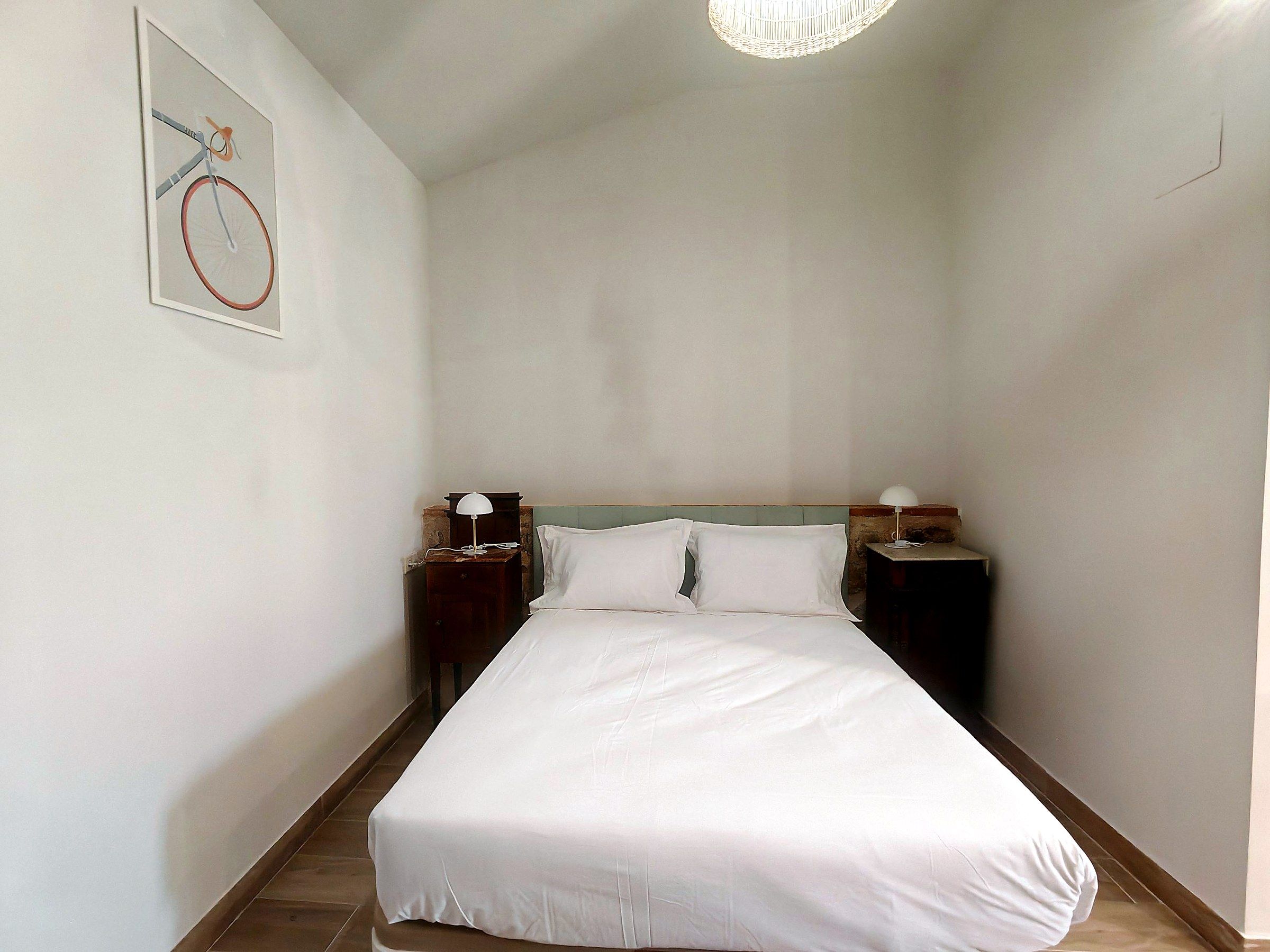 Habitación de hotel con una cama, dos mesitas de noche, una lámpara de pared y un cuadro enmarcado.