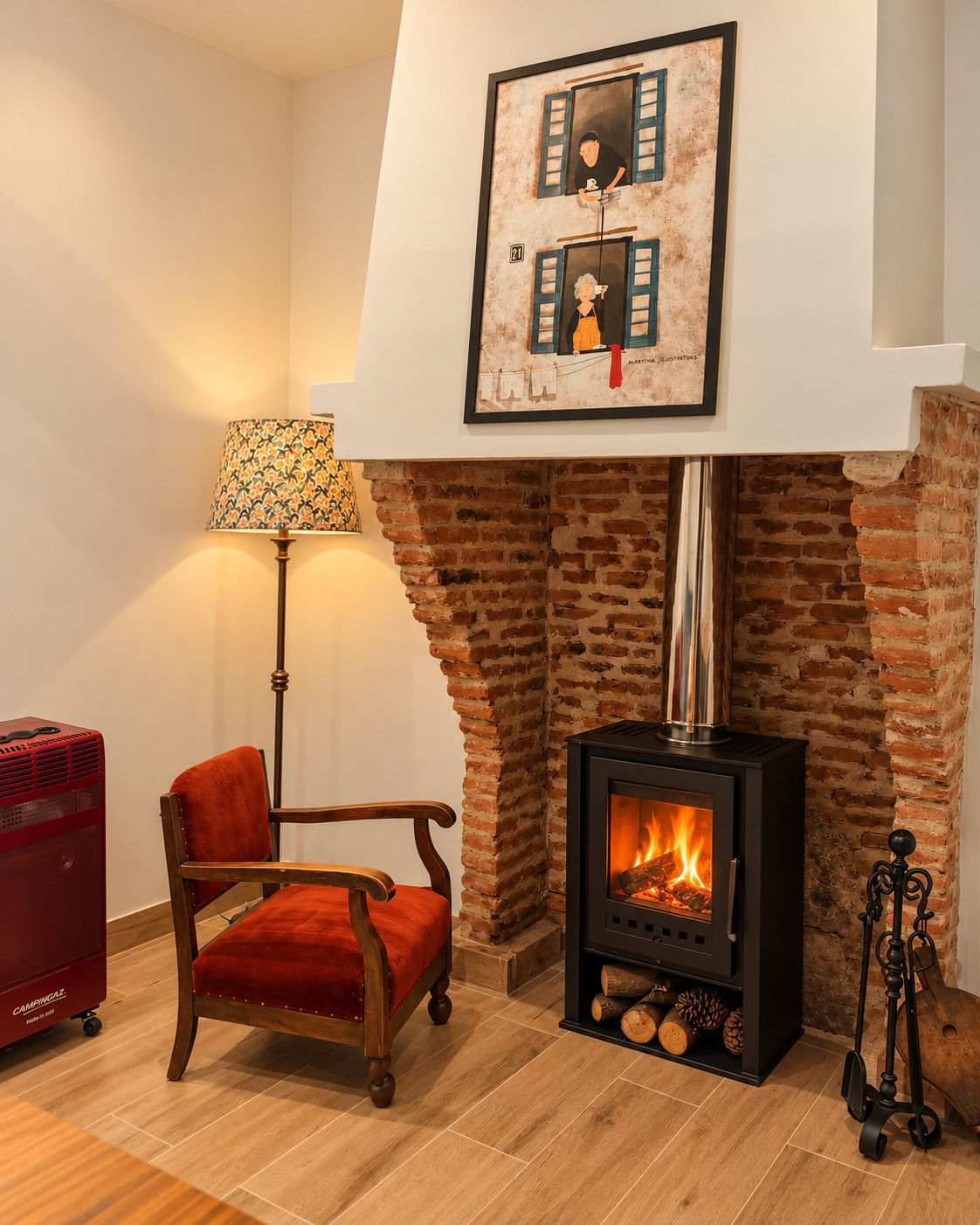 Una habitación acogedora con chimenea de ladrillo, un cuadro colgado encima, una silla roja, una lámpara de pie y un calefactor rojo.