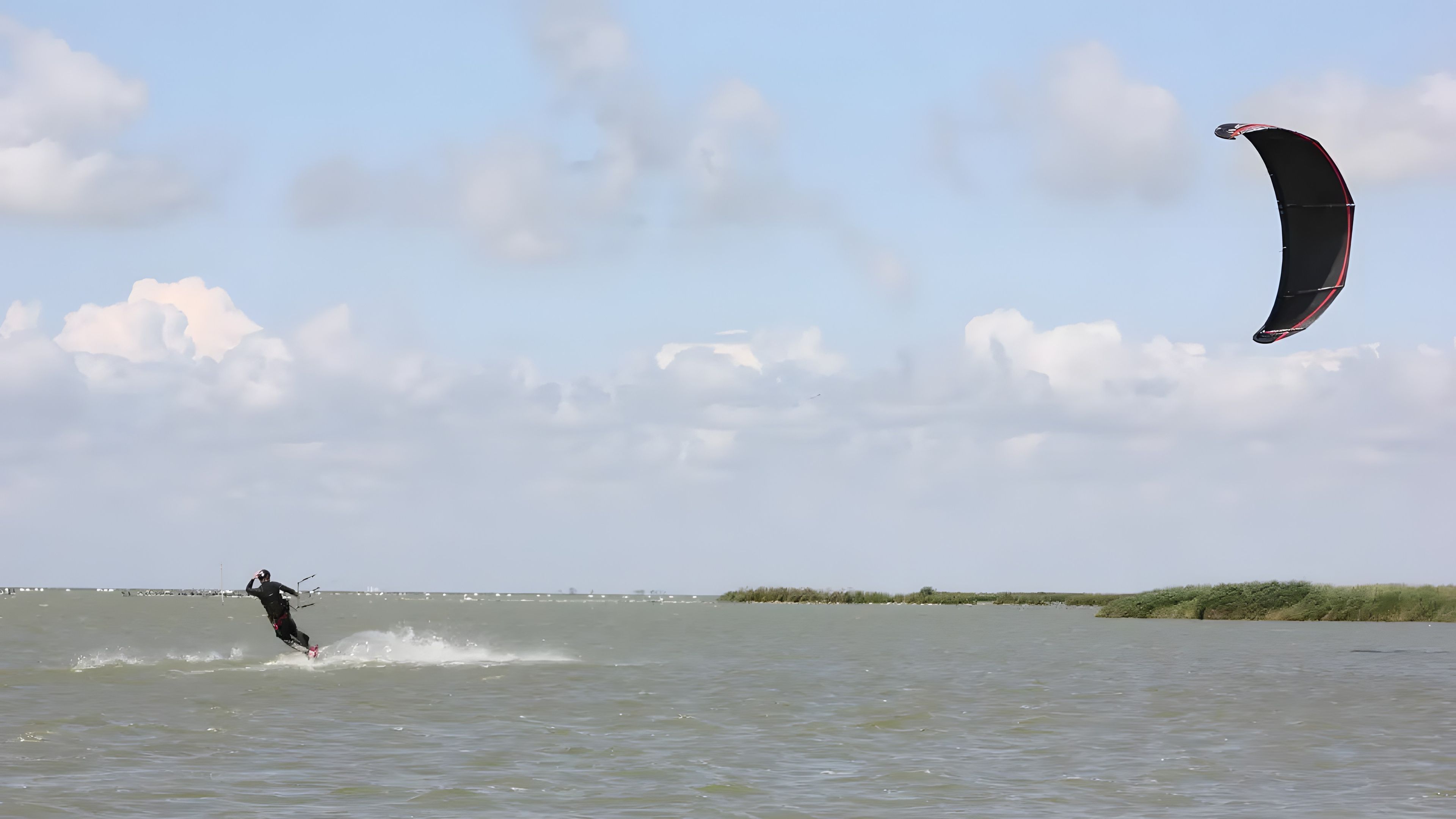 Una persona che pratica il kitesurf su un'area d'acqua, con cielo sereno e terra all'orizzonte.