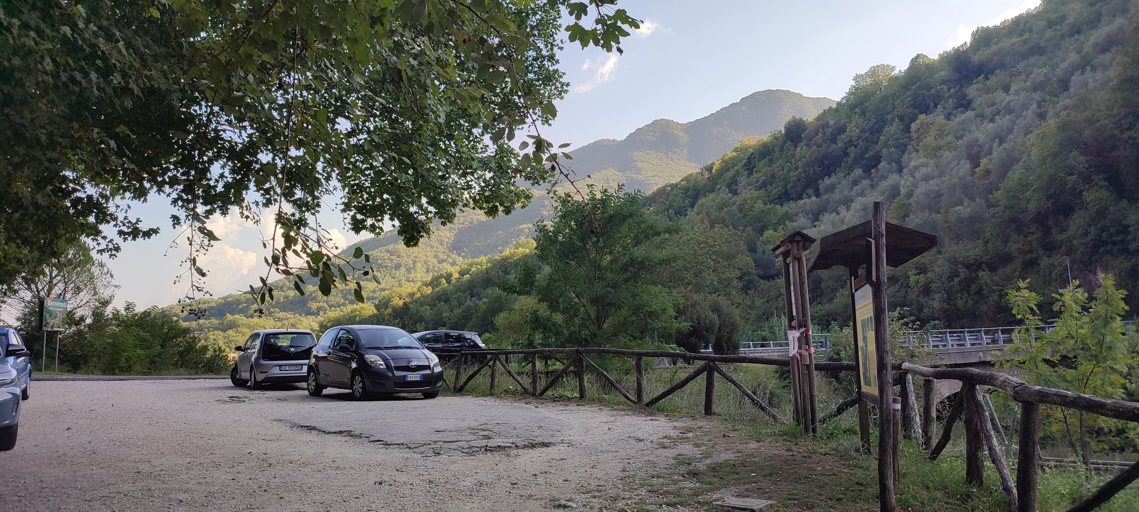 A gravel parking area with cars parked, surrounded by lush greenery and a mountainous backdrop.
