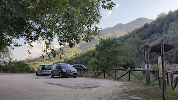 A gravel parking area with cars parked, surrounded by lush greenery and a mountainous backdrop.