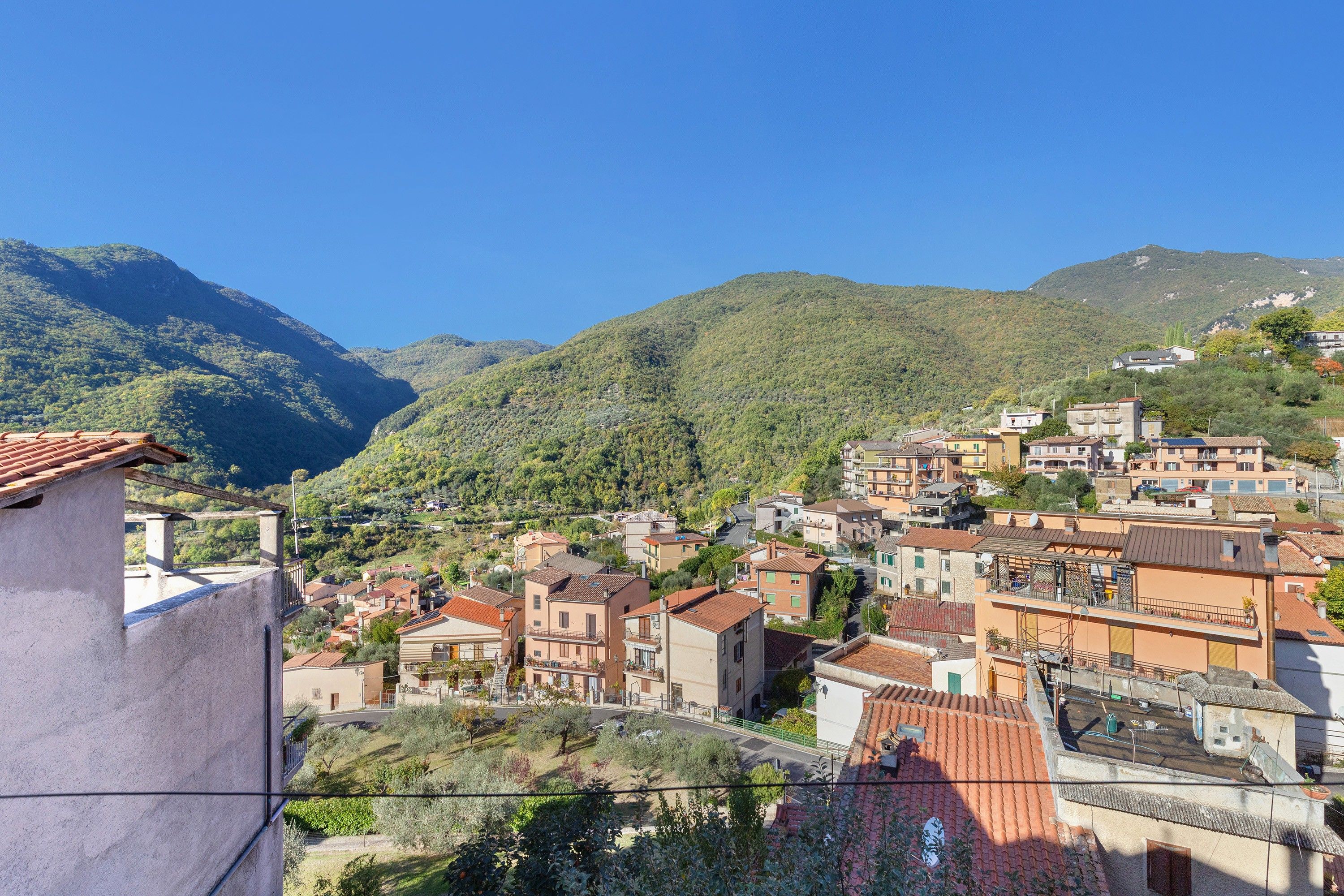 A hillside town with terracotta-roofed buildings, a clear blue sky, and lush green mountains in the background.