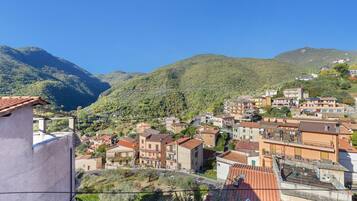 A hillside town with terracotta-roofed buildings, a clear blue sky, and lush green mountains in the background.