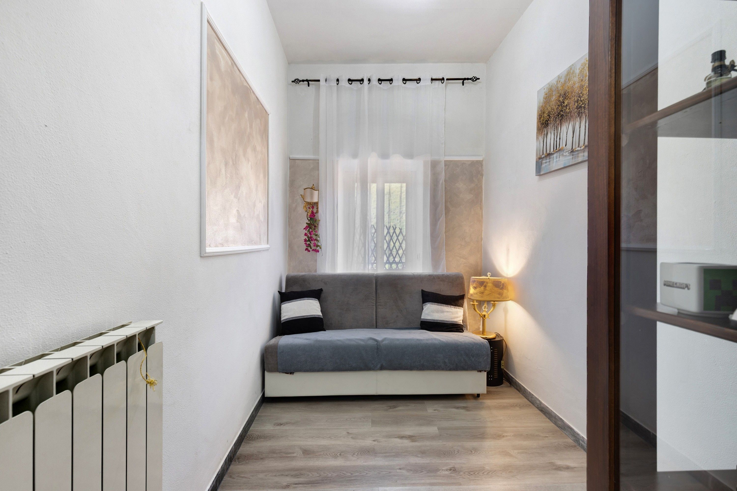 A narrow hallway with a grey sofa, a small table with a lamp, and a window with curtains.