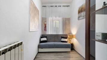 A narrow hallway with a grey sofa, a small table with a lamp, and a window with curtains.