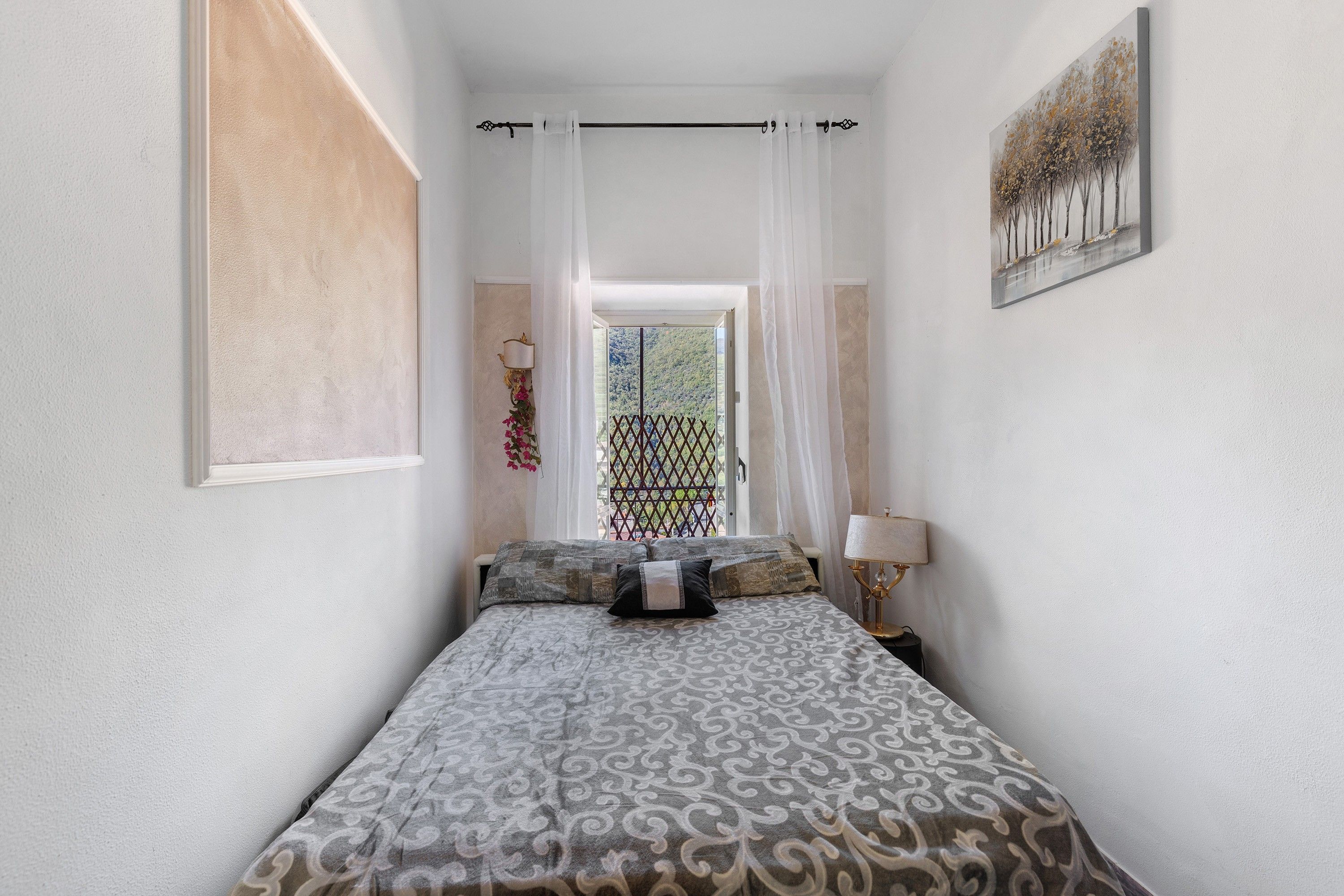 Een smalle hotelkamer met een bed, een raam met uitzicht op groen en een schilderij aan de muur.