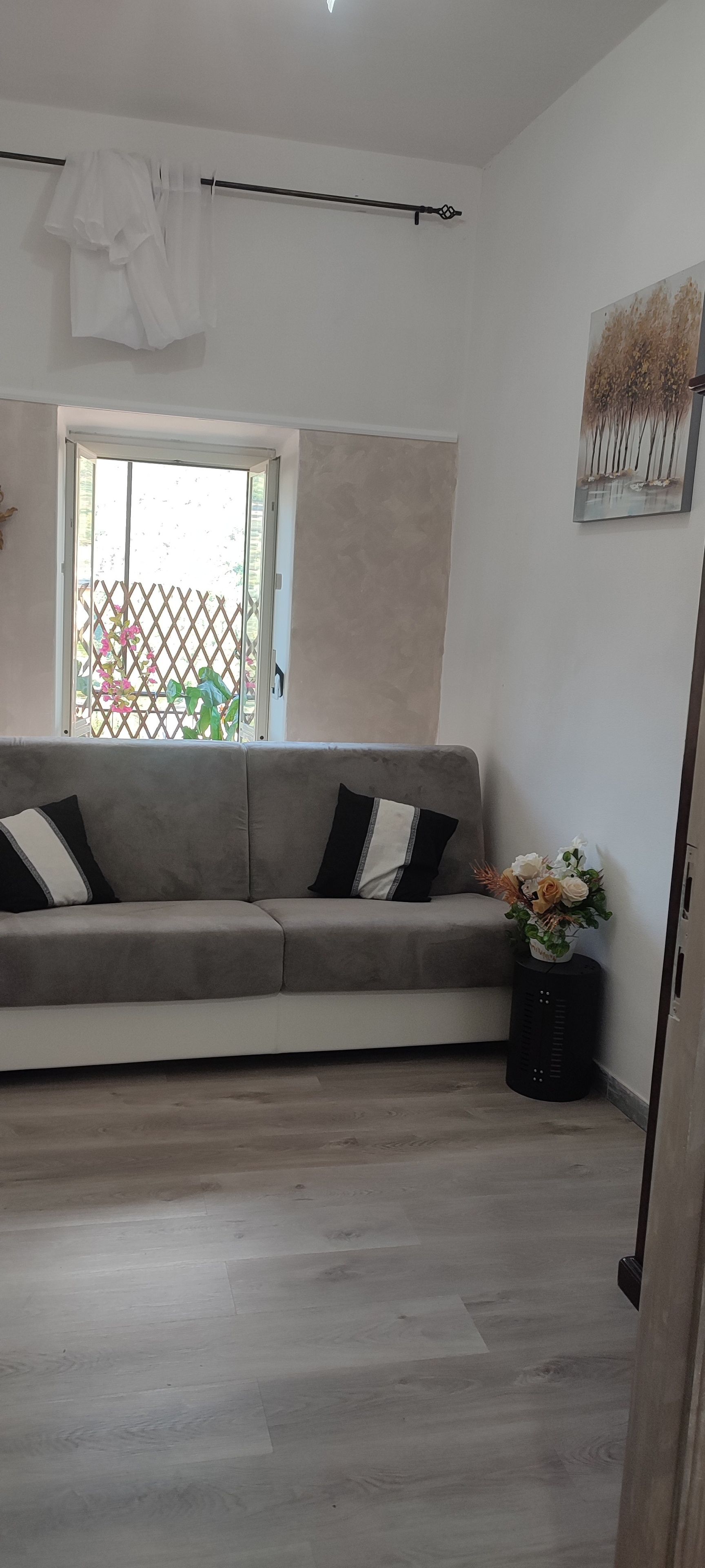 A living room with a grey sofa, a window with a view of greenery, and a wall-mounted artwork.