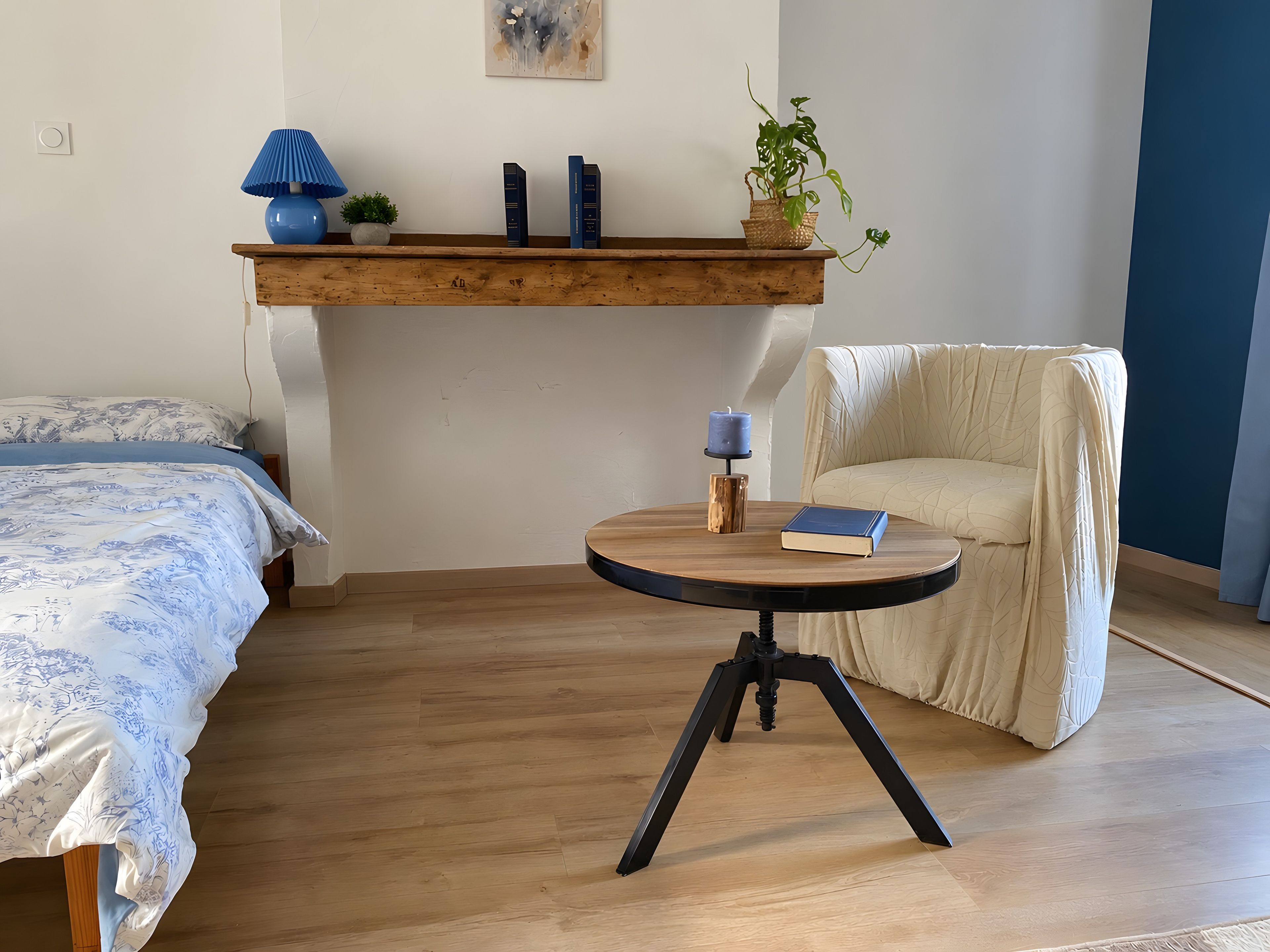 Une chambre à coucher avec un lit, une chaise, une petite table sur laquelle se trouvent un livre et une bougie, et une étagère avec des livres et une plante.