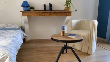 Une chambre à coucher avec un lit, une chaise, une petite table sur laquelle se trouvent un livre et une bougie, et une étagère avec des livres et une plante.