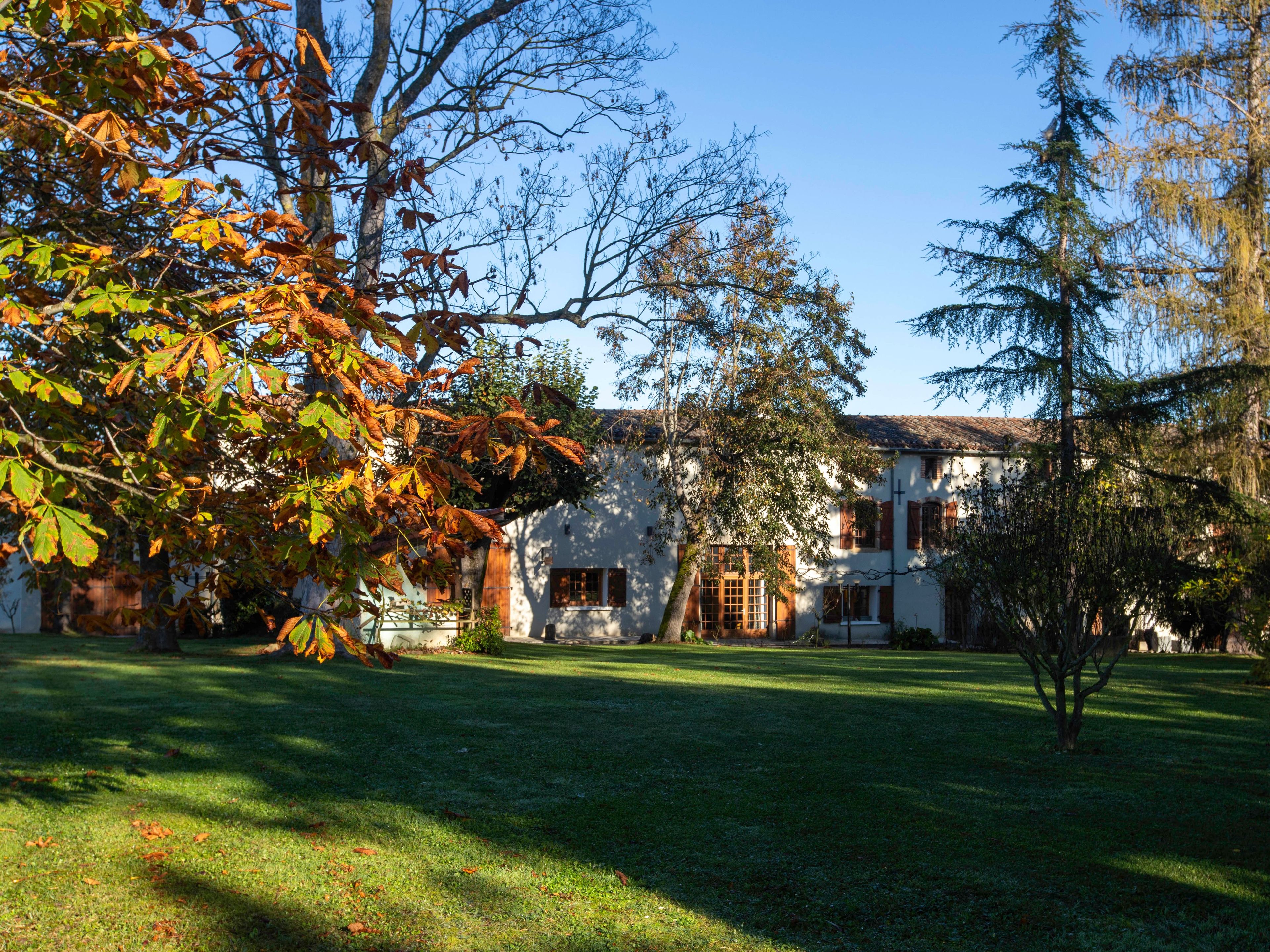 Un bâtiment traditionnel à la façade blanche et au toit recouvert de tuiles, entouré d’une pelouse bien entretenue et d’arbres aux feuilles d’automne.