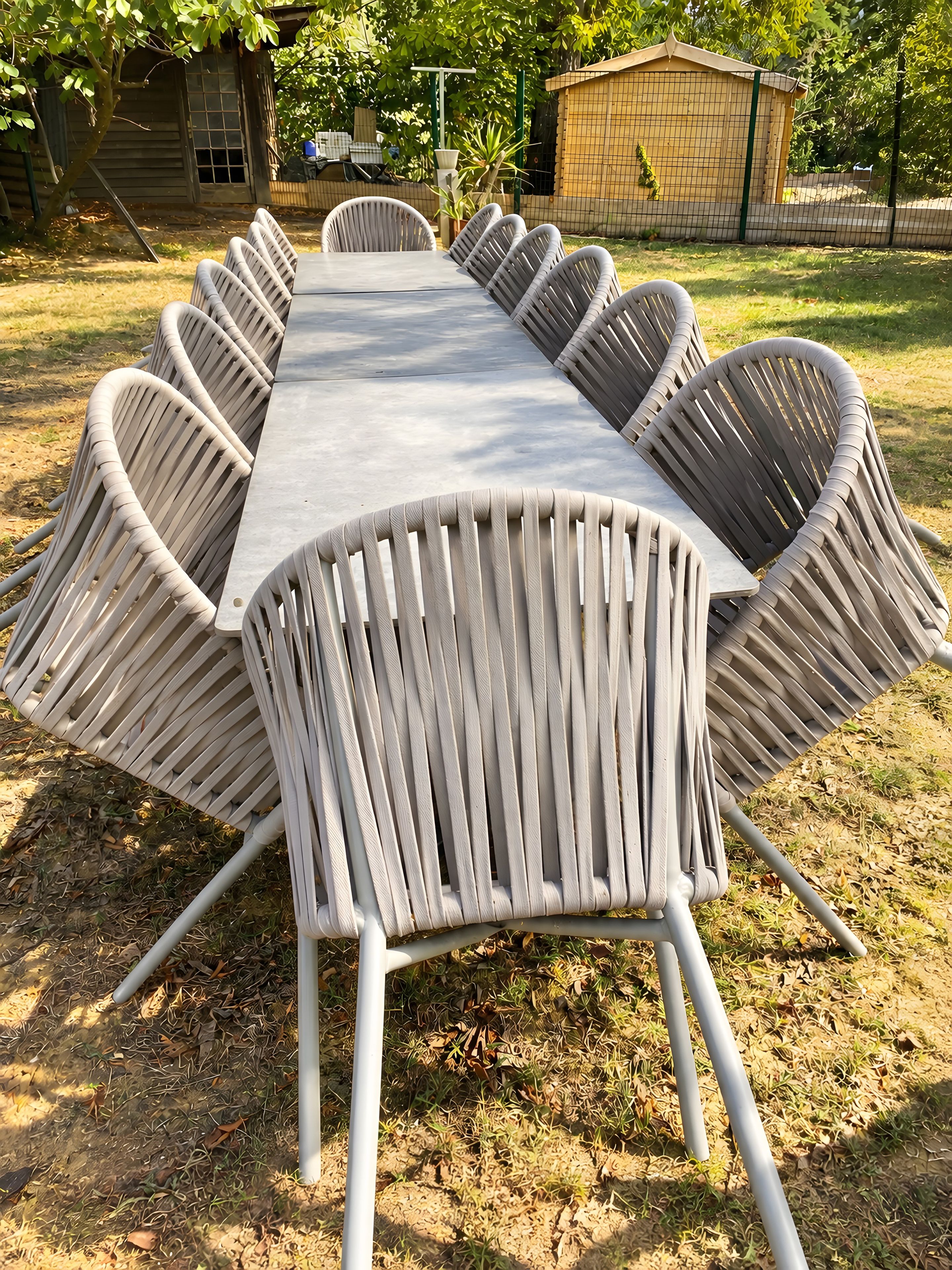 A rectangular outdoor table with multiple chairs, surrounded by grass and trees.