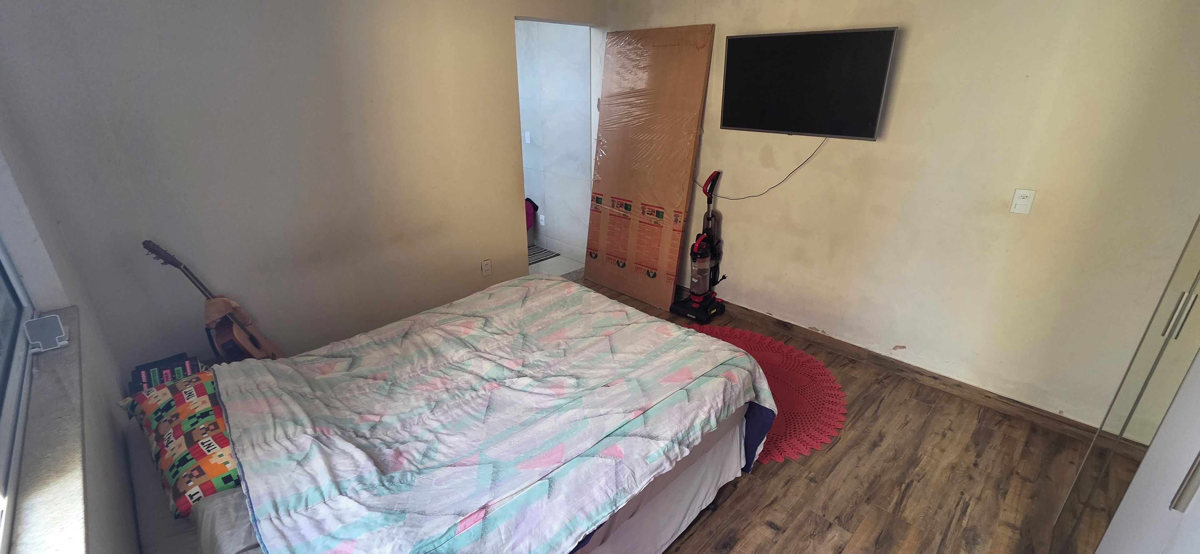 A bedroom with a bed, a guitar, a vacuum cleaner, and a TV on the wall.