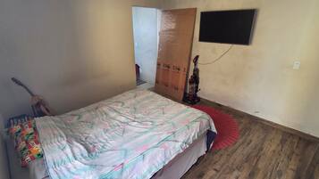 A bedroom with a bed, a guitar, a vacuum cleaner, and a TV on the wall.