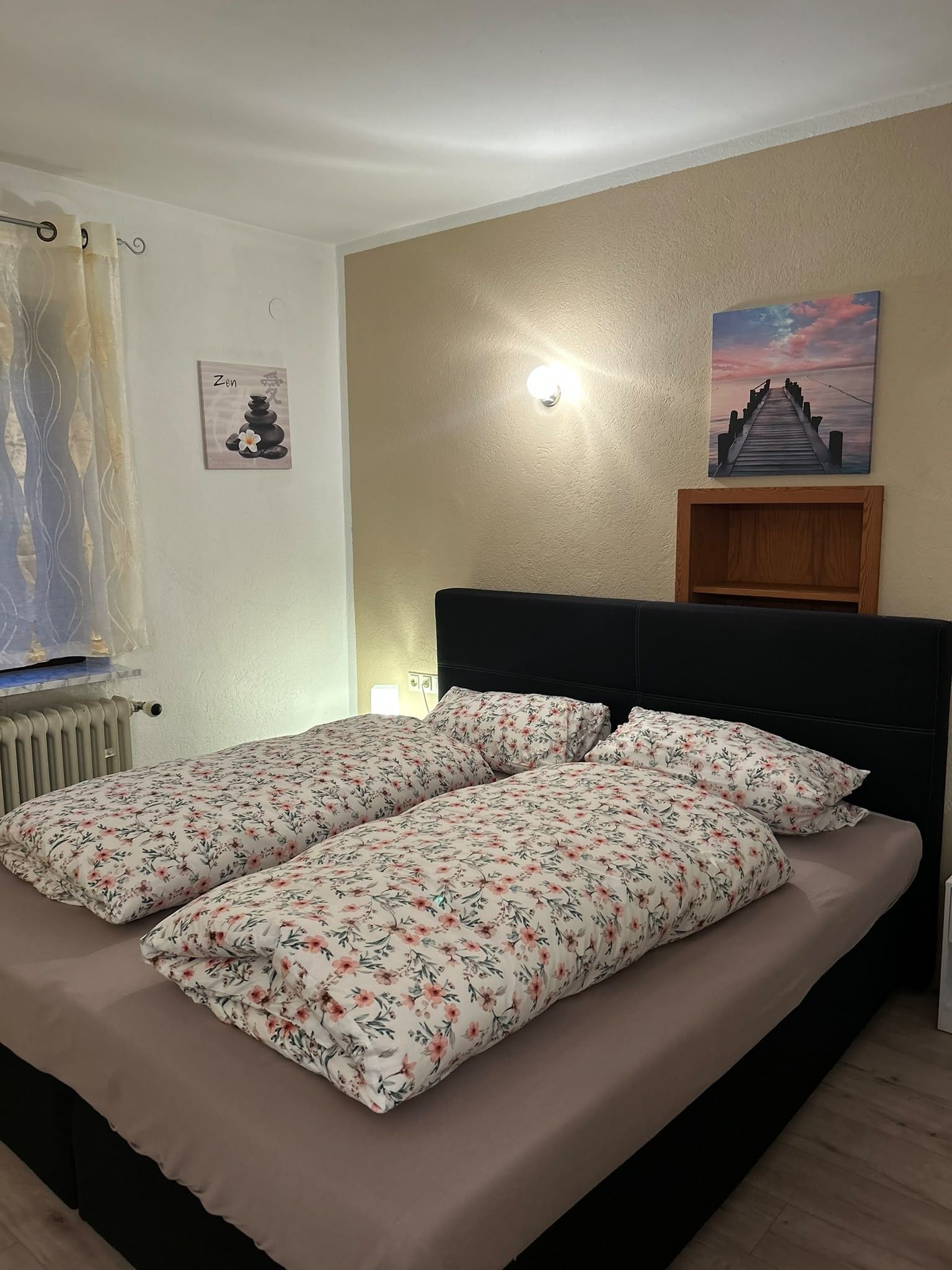 Un dormitorio con una cama, una lámpara de pared, un cuadro de un atardecer y un radiador.