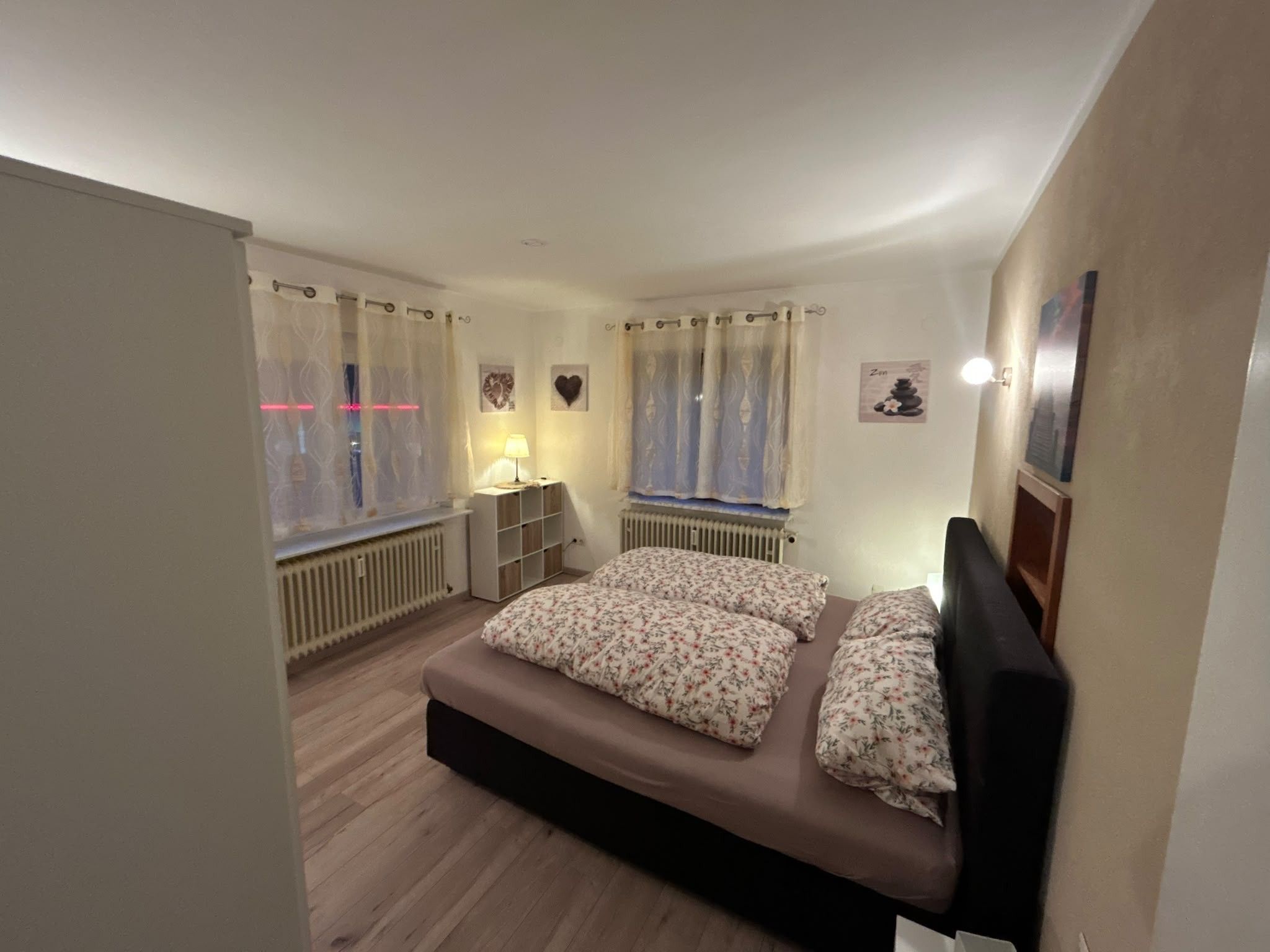 Une chambre à coucher avec un lit, deux fenêtres avec des rideaux, une table de chevet avec une lampe et un radiateur.