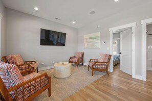 A spacious living room with hardwood flooring, a round ottoman, and two wooden chairs with patterned cushions. - Shore Thing: Oceanside, Private Pool, Hot Tub (Corolla)