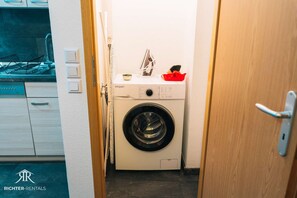 Ein kleiner Waschraum mit einer Waschmaschine mit Frontlader, einem Spülbecken und einer Tür zu einem Küchenbereich.