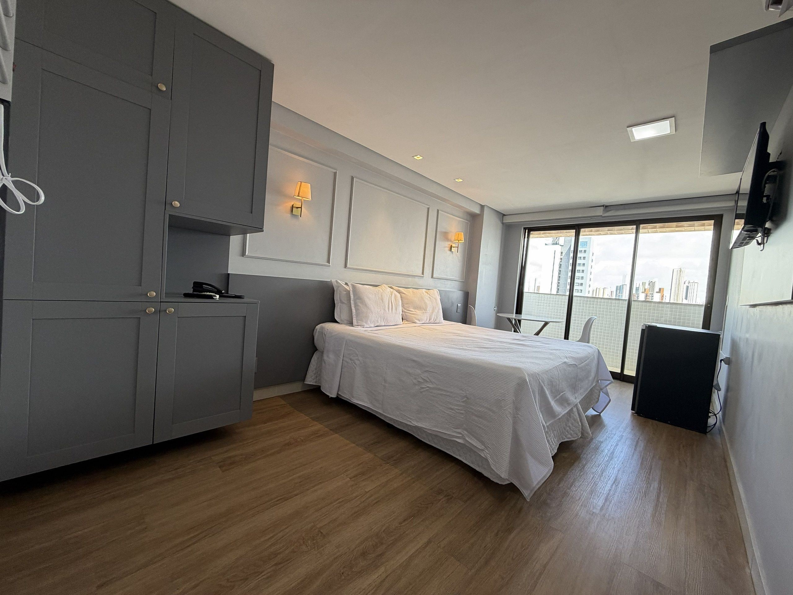 A hotel room with a bed, a desk, a chair, a balcony with a view of the city, and a TV mounted on the wall.