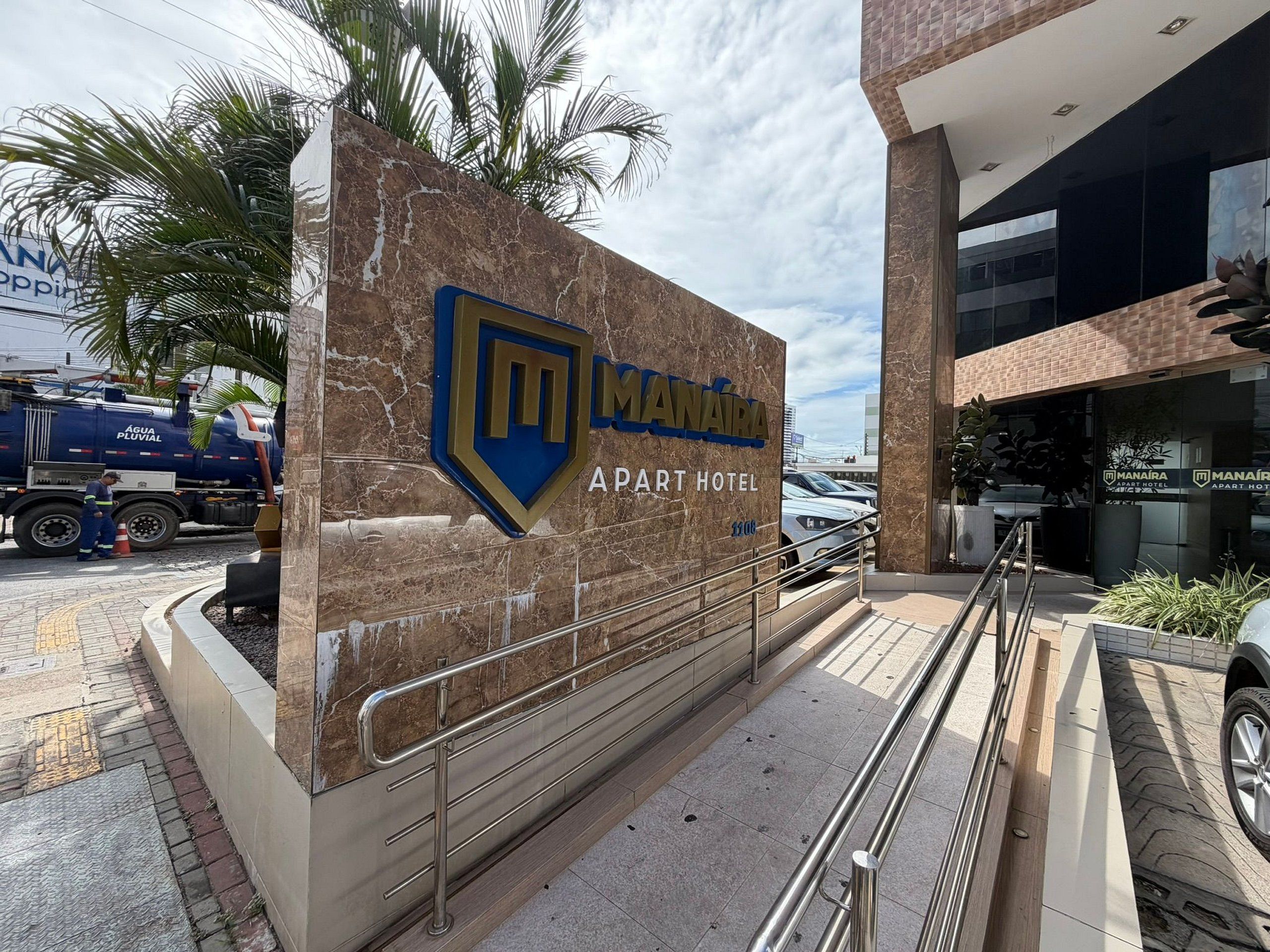 Entrada de hotel con fachada de mármol, escalera de metal y una palmera al fondo.