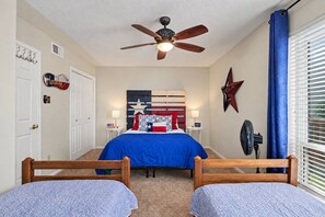 Ein Schlafzimmer mit Deckenventilator, einem Bett mit blauer Bettdecke, zwei Holzstühlen, einer Sterndekoration und einem Fenster mit Jalousien.