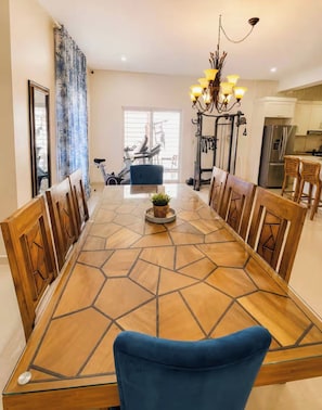 Un espace repas avec une longue table en bois, des chaises, un lustre et une vue sur une salle de sport.