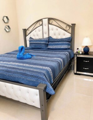 A bed with a tufted headboard, a blue patterned bedspread, and matching pillows. There is a blue towel on the bed, a black nightstand with a lamp, and a drawer unit. - Lovely House,pool,ac,near Sea,gym,bbq,puerto Silver (Montellano)