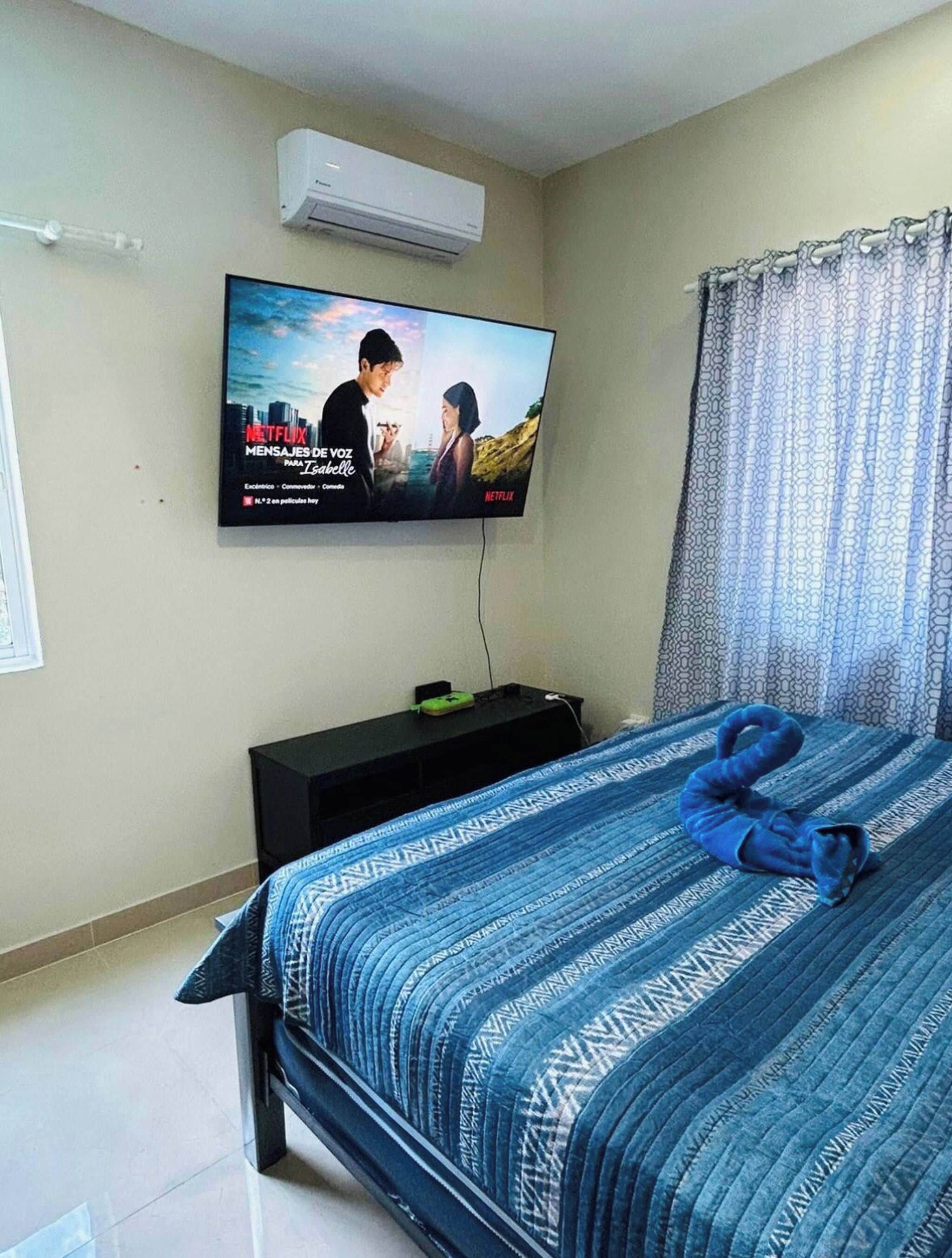 Une chambre à coucher avec un lit, un téléviseur fixé au mur, une table de chevet et une fenêtre avec des rideaux.