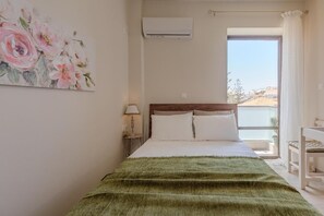 Quarto com cama, mesa de cabeceira, abajur, janela com vista e um quadro na parede.
