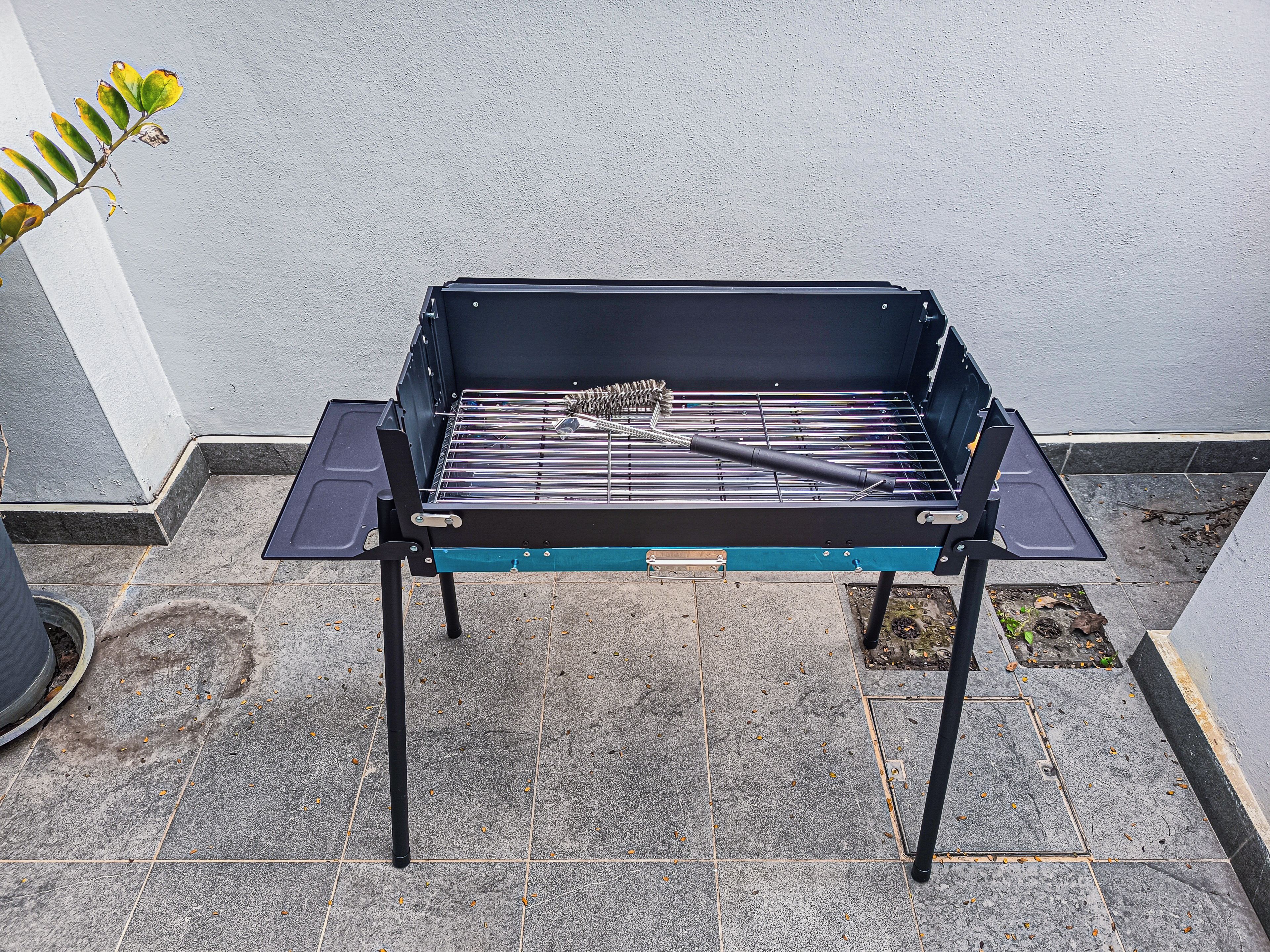 Una barbacoa amb una reixa metàl·lica i una broqueta, col·locada en un pati amb rajoles i una paret blanca al fons.
