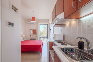 A compact kitchen with a red bed in the background, a window with a view, and a modern faucet. - Cozy Home Near The Sea 18 Studio with Balcony, Wi-Fi, and Air Conditioning (Ρέθυμνο)
