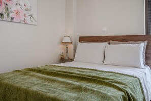 A bedroom with a wooden headboard, white bedding, a green blanket, a bedside lamp, and a floral painting on the wall. - Cozy Home Near The Sea 18 Studio with Balcony, Wi-Fi, and Air Conditioning (Ρέθυμνο)