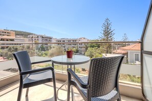 Un balcon avec des fauteuils en osier et une table, donnant sur un quartier résidentiel avec des immeubles et des arbres.