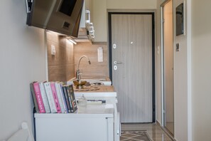 A compact kitchen with a built-in oven, a sink, and a microwave. There is a small countertop with a decorative bowl and a remote control. A door with a handle and a lock is visible in the background. - Cozy Home Near The Sea 19 Studio with Balcony, Wi-Fi, and Air Conditioning (Ρέθυμνο)