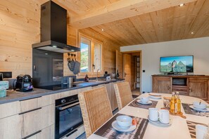 A modern kitchen with a built-in oven, a stove, and a large range hood. There is a dining area with a table set for two, and a television mounted on the wall. - Petit Chouda - Proche Télécabines (Saint-Chaffrey)