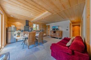 A spacious living area with a red sofa, a dining table with chairs, a kitchen area with a stove and refrigerator, and a television on the wall. - Petit Chouda - Proche Télécabines (Saint-Chaffrey)