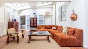 A living room with a large orange sofa, a glass coffee table, and a wooden chair. There is a bookshelf with red and white artwork, and a hat hanging on the wall. - Marais Saint Paul Atypique (Paris)