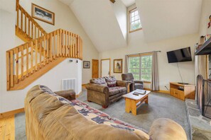 Ein geräumiges Wohnzimmer mit hoher Decke, einer Holztreppe, einem großen Fenster und einem an der Wand befestigten Flachbildfernseher.