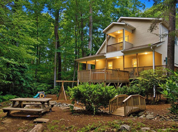 A two-story cabin with a deck, surrounded by dense forest. - Rushing Creek, EZ-Access, Hot Tub, Firepit, Playground (Maggie Valley)