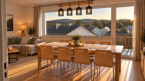 Uma sala de estar moderna com área de jantar, janelas amplas e vista para casas e colinas.